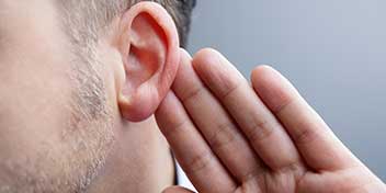 A close-up of a man’s ear with his hand cupped behind it, suggesting listening or hearing focus, set against a neutral gray background. No text or hearing aids are visible.