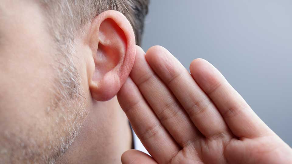 A close-up of a man’s ear with his hand cupped behind it, suggesting listening or hearing difficulty, set against a neutral gray background. No text is present.
