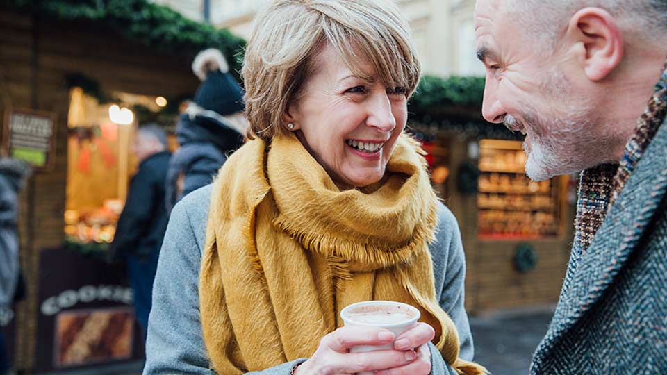 Couple sharing a conversation and enjoying a warm drink at an outdoor holiday market – 5 tips on starting the conversation about hearing loss.