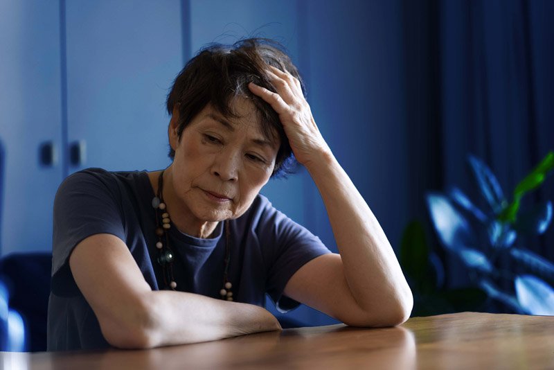 A person wearing a navy shirt and a beaded necklace rests their arm on a wooden table, holding their head thoughtfully in a softly lit room with blue decor and indoor plants nearby.