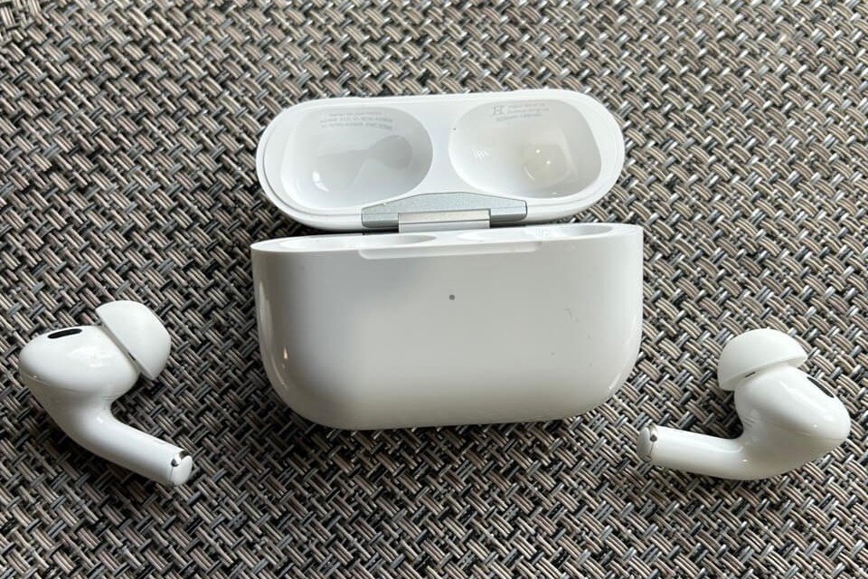White wireless earbuds rest beside an open charging case on a woven textured surface. The case lid is flipped upward, revealing interior dock spaces.
