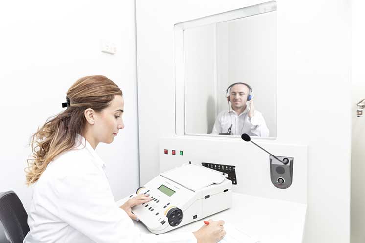 Man undergoing a hearing test in a soundproof booth while a hearing specialist operates testing equipment.