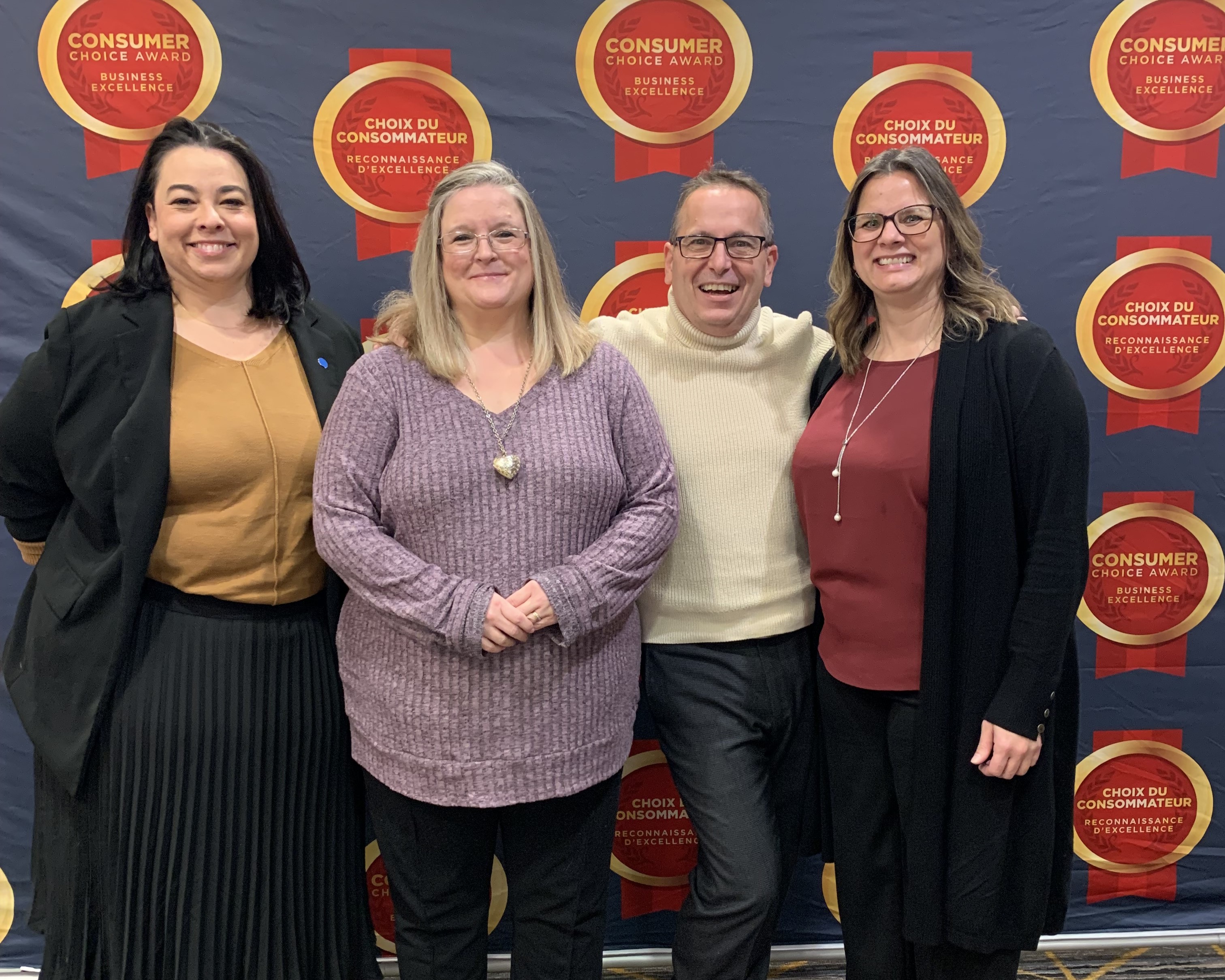 Four individuals stand closely together in front of a backdrop displaying ’Consumer Choice Award Business Excellence’ and ’Choix du Consommateur Reconnaissance d’Excellence’ logos, suggesting an award or recognition event.