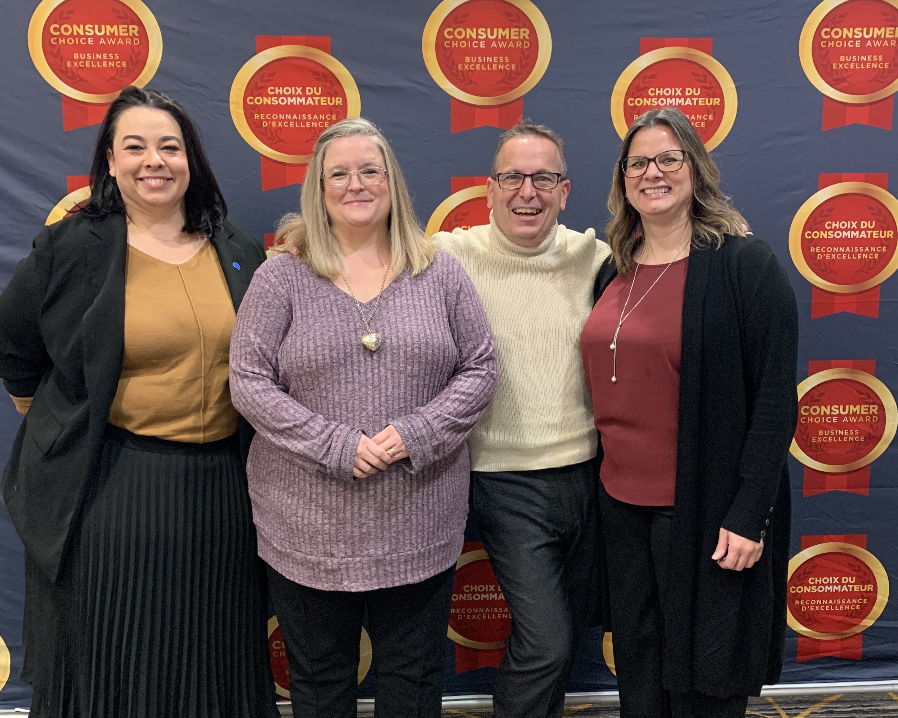 Four individuals stand closely together in front of a backdrop displaying ’Consumer Choice Award Business Excellence’ and ’Choix du Consommateur Reconnaissance d’Excellence’ logos, suggesting an award or recognition event.