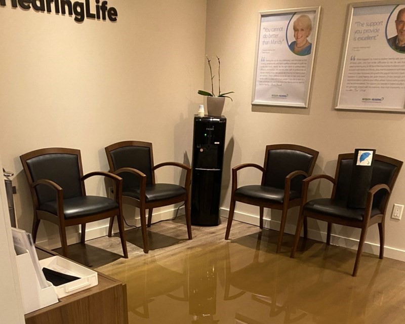 Four wooden chairs with black cushions are arranged in a waiting area at a HearingLife hearing clinic. A black water dispenser, a small plant, and framed wall posters displaying text, including client testimonials, complete the room’s minimalist design. The posters show HearingLife’s logo at the bottom, enhancing the hearing care clinic’s branding.