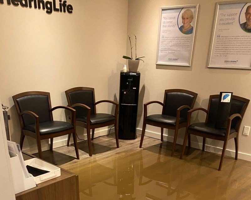 Four wooden chairs with black cushions are arranged in a waiting area at a HearingLife hearing clinic. A black water dispenser, a small plant, and framed wall posters displaying text, including client testimonials, complete the room’s minimalist design. The posters show HearingLife’s logo at the bottom, enhancing the hearing care clinic’s branding.