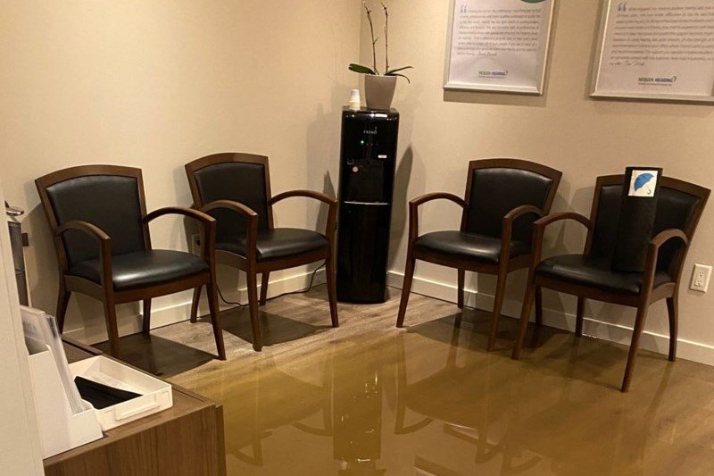 Four wooden chairs with black leather upholstery surround a black water dispenser in a small waiting area with light beige walls. Framed HearingLife testimonials are displayed on the wall above.