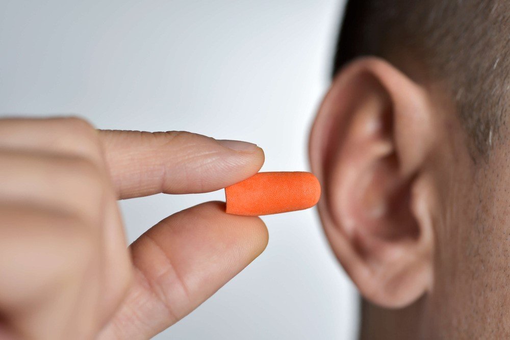 A hand grips an orange foam earplug near an ear, poised for insertion. The background is softly blurred, emphasizing hearing protection and ear care in a personal health context.