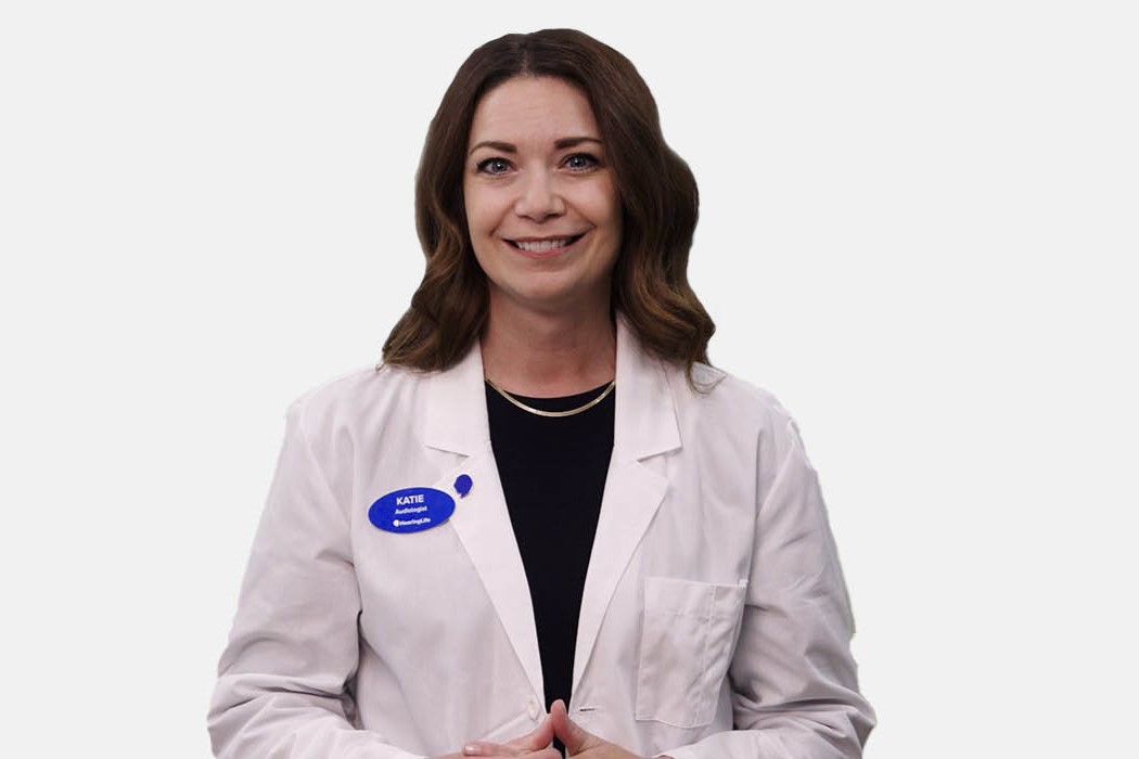 A professional audiologist, identified as ’Katie’ by a blue HearingLife name tag, wears a white lab coat over dark clothing against a plain white background, conveying expertise in hearing care services.