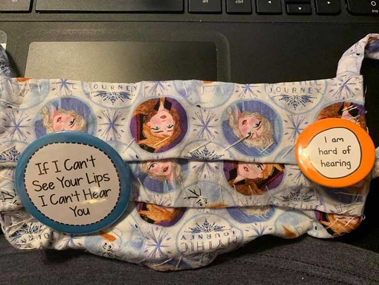 Decorative face mask with Frozen character designs, featuring buttons that read ’If I can’t see your lips, I can’t hear you’ and ’I am hard of hearing,’ placed on a laptop keyboard.