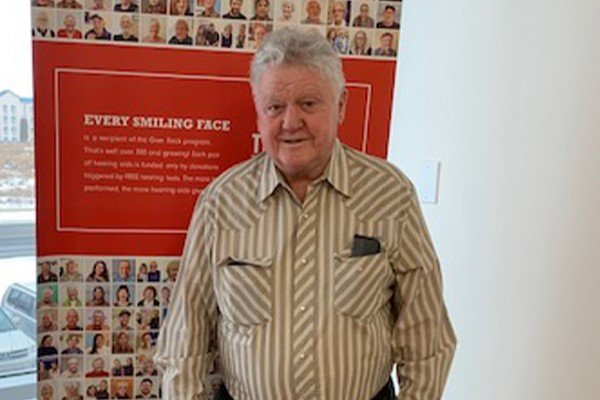 CFBH program recipient standing in front of a red banner showcasing success stories of hearing care improvements.