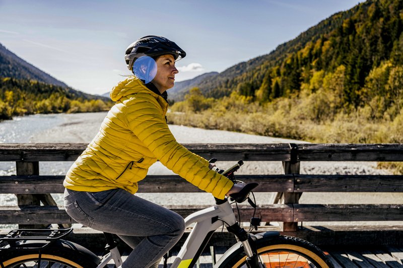 A person cycling on a scenic mountain trail, wearing a yellow jacket, helmet, and visible hearing protection near a wooden bridge over a river.