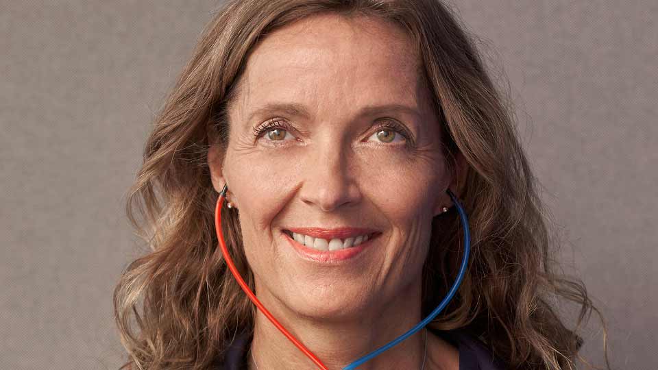 A person with partially visible long, wavy hair wears a blue and red binaural stethoscope around their neck against a neutral, textured background, emphasizing hearing health equipment in a clinical setting.