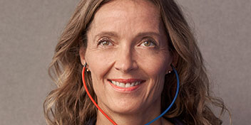 A person with shoulder-length wavy hair, blurred face, wears red and blue tubing resembling binaural audiometric headphones, used during hearing tests in a clinic setting. No text is visible.