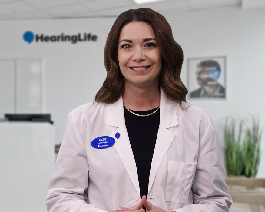 A female audiologist, wearing a white lab coat with a visible name tag reading ’KATIE, Audiologist @HearingLife,’ stands in a modern hearing clinic branded with a ’HearingLife’ logo in the background.