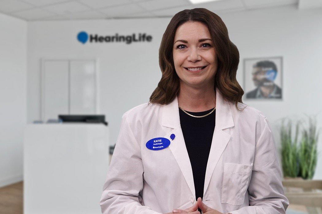 A woman in a white lab coat, wearing a blue name tag reading ’KATIE, Audiologist, @HearingLife,’ stands in a modern hearing clinic. The HearingLife logo is visible on the white wall in the background alongside a framed photo and indoor plants.