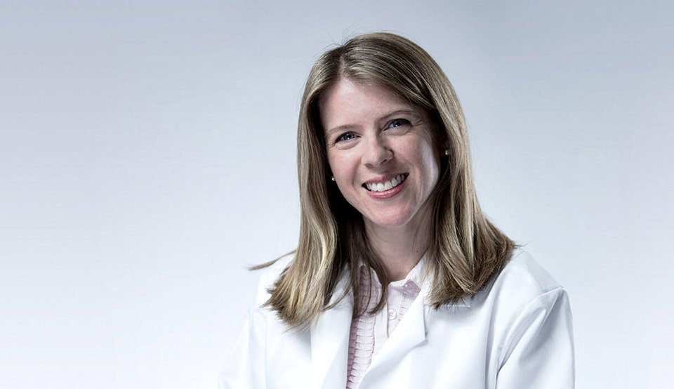 A woman wearing a white lab coat and light pink shirt sits against a neutral gray background, representing a professional environment like a hearing clinic associated with hearing care services. No visible text or branding.