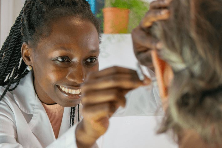 A hearing care professional adjusts a hearing aid for a patient with gray hair. The setting appears to be a hearing clinic with a white coat and indoor greenery visible.