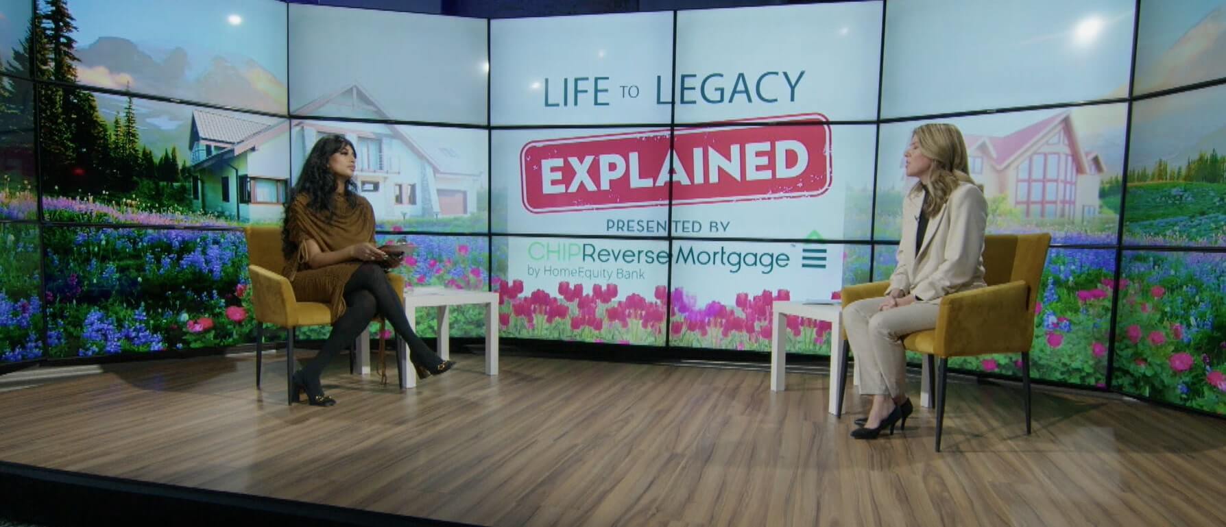 Two women sit in mustard-yellow chairs on a wooden studio floor, discussing ’LIFE TO LEGACY EXPLAINED’ (text displayed on a large screen). The sponsor, ’CHIP Reverse Mortgage by HomeEquity Bank,’ is also prominently displayed. The screen background depicts a colorful garden with homes and mountains.
