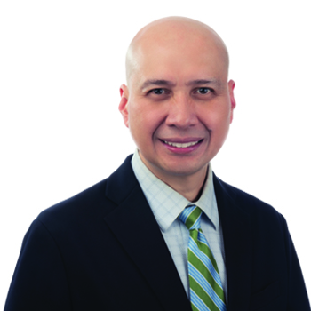A person wearing a dark blazer, light checkered shirt, and green striped tie stands against a white background. Their face is intentionally blurred to obscure identity. No text is present.