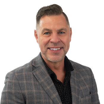 A man wearing a blue plaid blazer and black dotted shirt stands against a plain white background. The face is intentionally blurred, providing no identifiable features. No text is present.