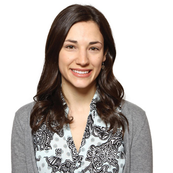 A woman with shoulder-length, wavy dark brown hair is wearing a patterned blouse and gray cardigan. Her face is intentionally blurred. The background is plain white, offering a clinical or professional setting suitable for a hearing care context. No text or visible hearing aids are shown.