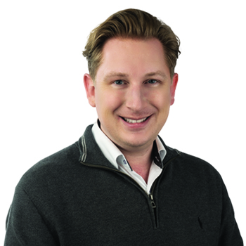 A person with short, light brown hair wearing a dark grey quarter-zip sweater over a white collared shirt is posed against a plain white background. The face is blurred.