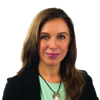 A professional woman with shoulder-length brown hair wears a black blazer, a mint-green top, and a circular pendant necklace, posing against a plain white background in a polished setting. No text visible.
