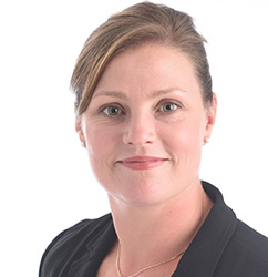 A professional wearing a black blazer and a gold chain necklace is positioned against a plain white background. The face is intentionally blurred, focusing on attire and setting clarity. No visible text or hearing aids are present.