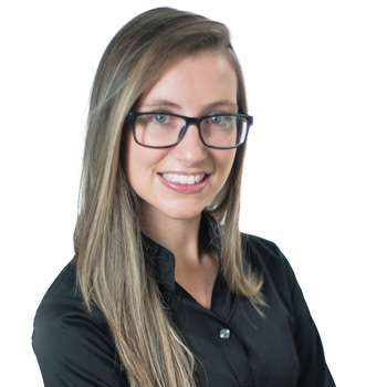 A person with long, straight brown hair wearing glasses and a black collared shirt stands confidently against a white background. Their face is intentionally blurred for privacy. No text is visible.