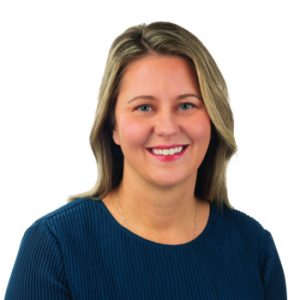 A person with shoulder-length blonde-brown hair wears a textured navy blue top. Their face is blurred for anonymity, set against a plain white background commonly used in professional profiles or hearing clinic contexts.