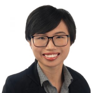 A person with short, dark hair wearing a collared shirt and a black blazer, set against a white background. The face is blurred, obscuring facial features completely.