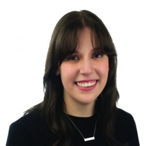 A person with long dark hair, wearing a black top and a rectangular silver necklace, is set against a plain white background. The face is intentionally blurred. No text is visible.