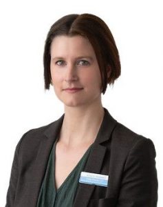 A professionally dressed individual wears a dark blazer over a green top, accompanied by a name badge featuring a blue and white design, likely in a healthcare or hearing clinic setting.