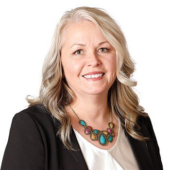 A professionally dressed individual wearing a black blazer, white blouse, and a statement necklace with colorful stones stands against a white background, representing a staff member from a hearing clinic.