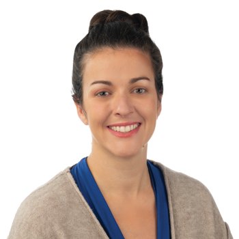 A person with dark hair styled in a bun wears a beige cardigan over a blue top. The face is intentionally blurred, and the background is entirely white.