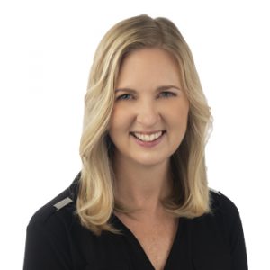 A woman with shoulder-length blonde hair wears a black shirt, standing against a plain white background. Her face is blurred, making facial details indistinguishable. No hearing aids or text are visible.