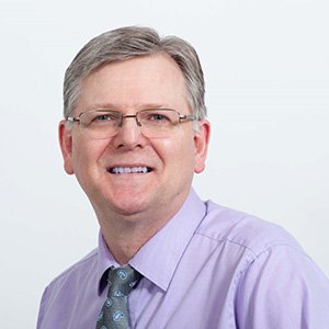 A person in a light purple dress shirt and patterned tie is positioned against a plain white background. The facial area is blurred, and no identifiable text or hearing aid branding is present.