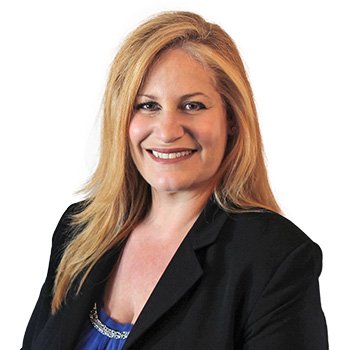 A professional woman with long, blonde hair wears a black blazer over a blue top with beaded details. The background is plain white, suggesting a corporate or hearing care clinic setting. No visible text or hearing aids.
