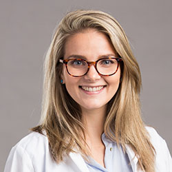 A professional wearing a white coat, likely an audiologist, stands against a plain gray background. The outfit suggests a hearing clinic setting, possibly related to hearing tests or hearing aid consultations.