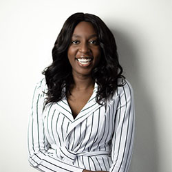 A woman wearing a white, striped blazer stands with arms crossed against a plain white background. Her face is intentionally blurred, emphasizing her attire and posture.