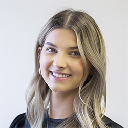 A woman with shoulder-length blonde hair wears a discreet, behind-the-ear hearing aid, positioned in a neutral indoor setting with a plain light-colored background. No visible text appears in the image.