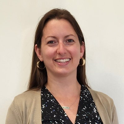 A woman with long brown hair, wearing gold hoop earrings, a black patterned blouse, and a beige cardigan, stands against a plain, light-colored wall. The face is intentionally blurred. No text or hearing aid branding is visible.