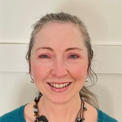 A person with grayish-brown hair tied back is wearing a teal top and a beaded necklace. The background features a plain, off-white wall. The face is intentionally blurred. No text or hearing aid branding is visible.