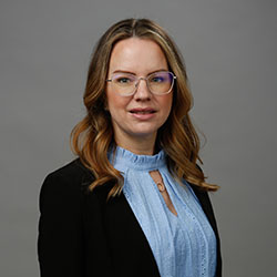 A professionally dressed individual wearing a black blazer over a light blue blouse stands in front of a neutral gray background. The face is intentionally blurred, anonymizing the subject.