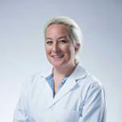 A professional wearing a white lab coat stands against a neutral background, likely representing an audiologist or hearing specialist in a hearing clinic setting. No text is visible.
