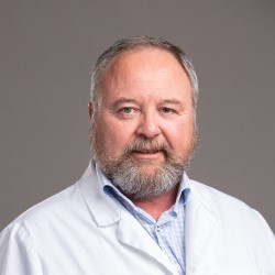 A professional wearing a white lab coat over a light blue patterned shirt, standing against a plain gray background, suggesting a clinical or hearing care setting. No visible text or branding.