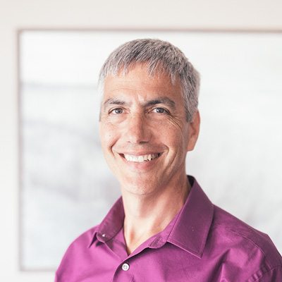 A middle-aged man with salt-and-pepper hair wearing a magenta dress shirt stands indoors against a blurred white and gray background, potentially in a professional setting like a hearing clinic. No text visible.