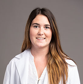 A professional in a white lab coat stands against a neutral gray background. The blurred face obscures identity, suggesting representation of a medical or audiology expert, possibly in a hearing clinic setting. No text is visible.