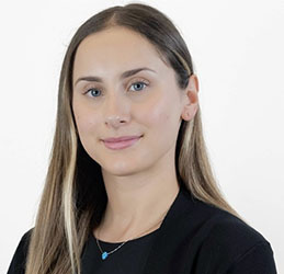 A person with long, light-brown hair wears a black top and a delicate necklace with a small blue pendant, standing against a plain white background. No text or hearing aids visible.
